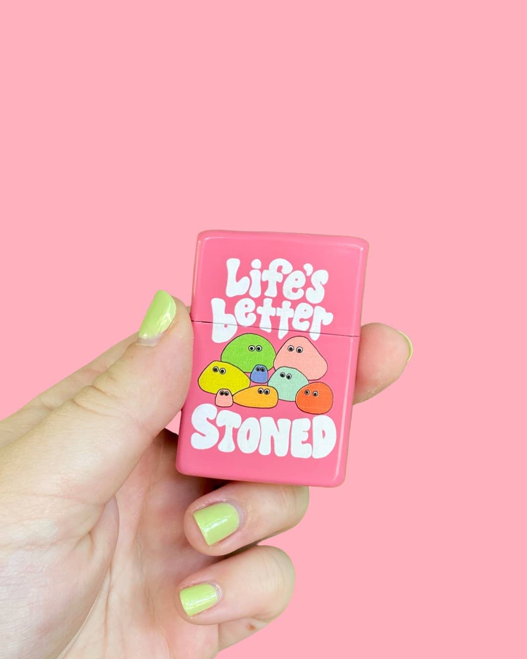 A hand with pastel yellow-green nail polish holds The Peach Fuzz Lifes Better Stoned Lighter, a pink, eco-friendly, refillable lighter featuring cartoon rocks and text, set against a solid pink background.
